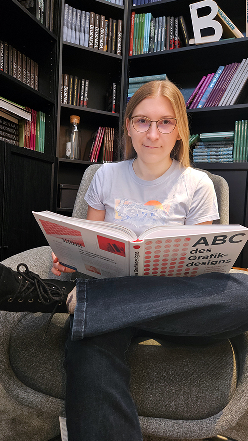 Andrea Maria Pauritsch Medienfachfrau beim Lesen eines Grafikdesign Buchs vor einem Bücherregal